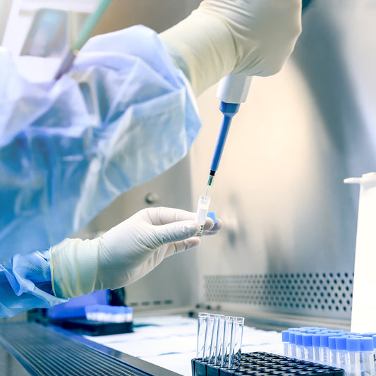 Person in a lab setting using a pipette with test tubes and lab equipment in the background.